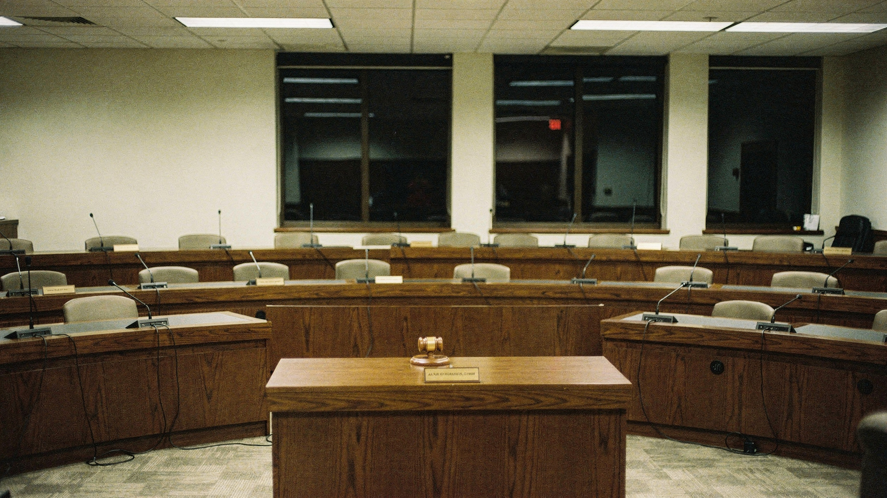 A documentary photograph of an empty hearing room, long wooden council desks in curved rows with microphones and nameplates, tall windows dark beyond, fluorescent light pooled on the carpet