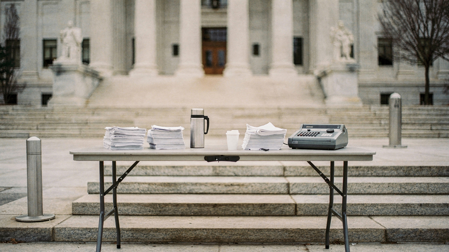 A folding table outside a federal courthouse with paper stacks, a coffee thermos, and a stenographer's machine, morning light