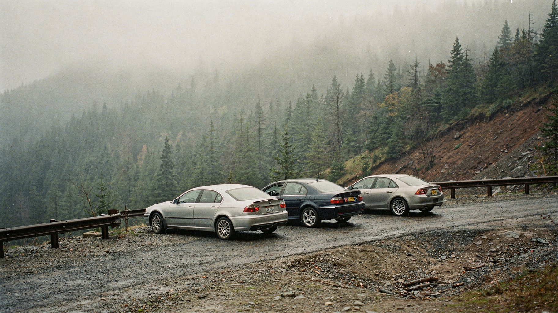 A documentary photograph of a mountain switchback road in early morning fog, a line of four cars from different manufacturers parked nose to tail at a gravel turnout, brake lights off, nobody in frame