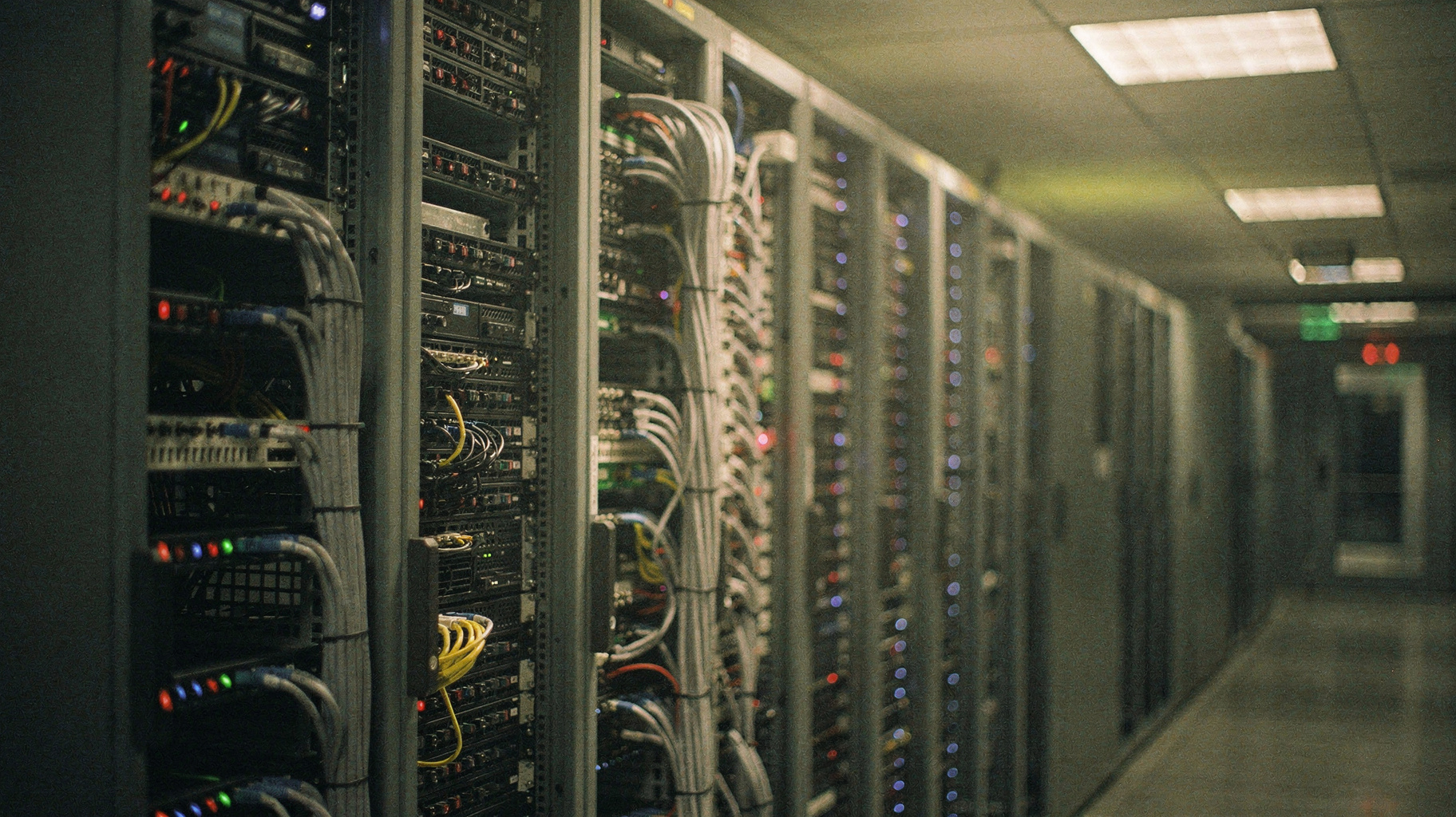 A rack of server boards in a chilled data-center aisle, dense cabling combed down one side, indicator lights lit, cold overhead light, photographed from the end of the row at a documentary distance