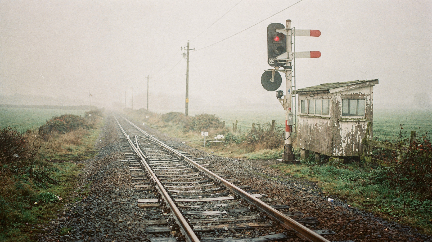 A documentary photograph of a rural railway semaphore signal set at caution in morning fog, telephone poles receding down the line, wet gravel ballast, nobody in frame