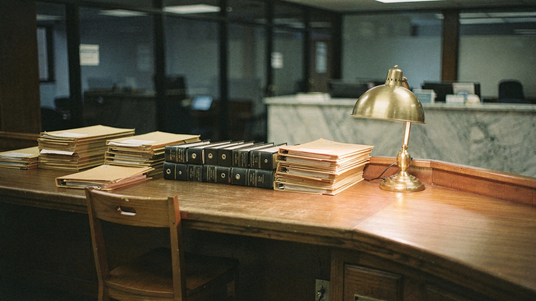 A documentary photograph of a quiet bank lending office after hours, document folders stacked on a long wooden counter, one desk lamp lit