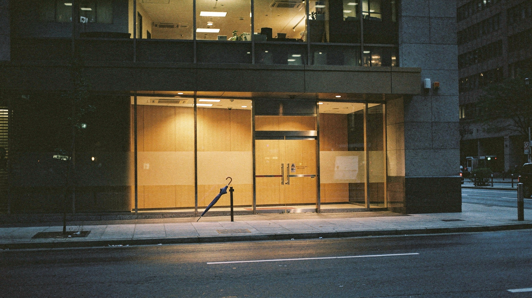 A documentary photograph of a lit but empty office building lobby at night, seen from across the street, a closed umbrella resting against the glass door