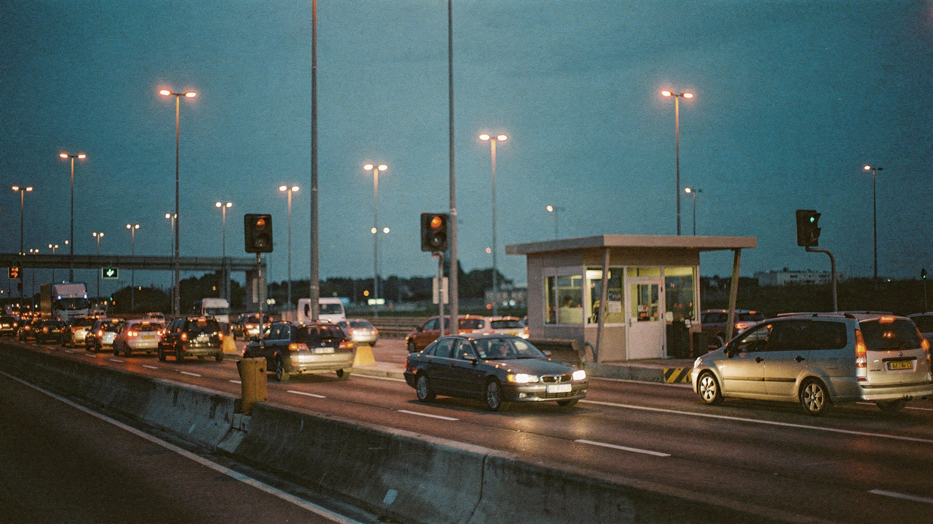 A documentary photograph of a highway toll plaza at dusk with one attended booth open, a single sedan passing through, sodium lamps glowing against a deep blue sky