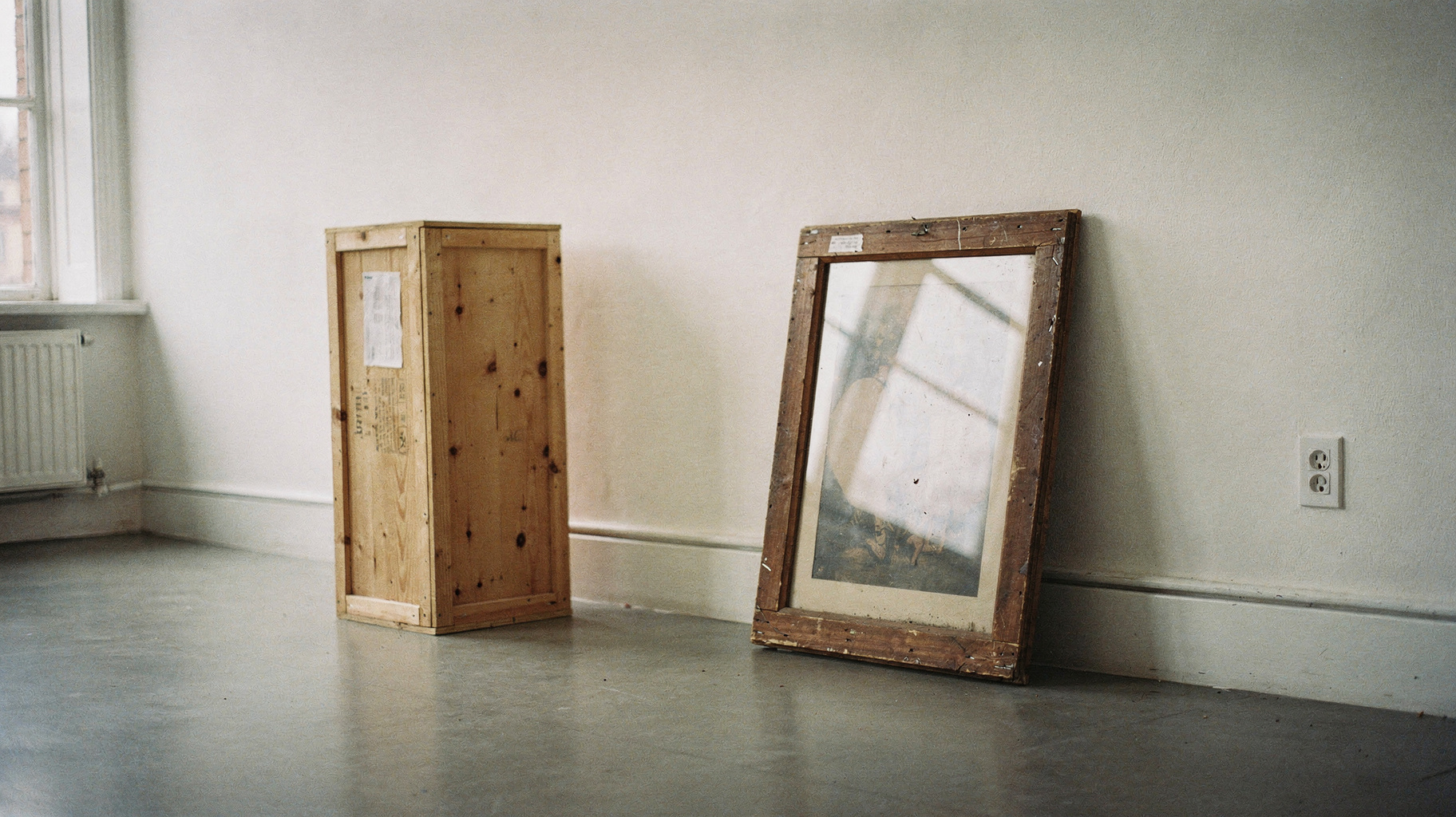 An old framed picture leaning against a bare gallery wall, a blank wooden shipping crate standing beside it on the floor, soft window light from the left