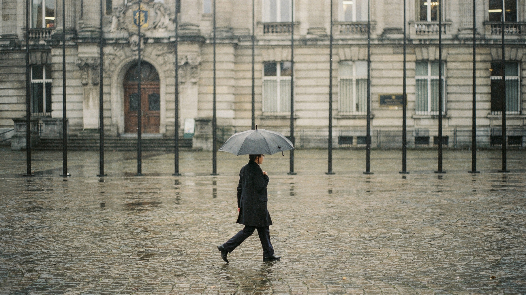 A wide Brussels government plaza in soft rain, a lone official crossing with an umbrella, a row of black flag poles without flags