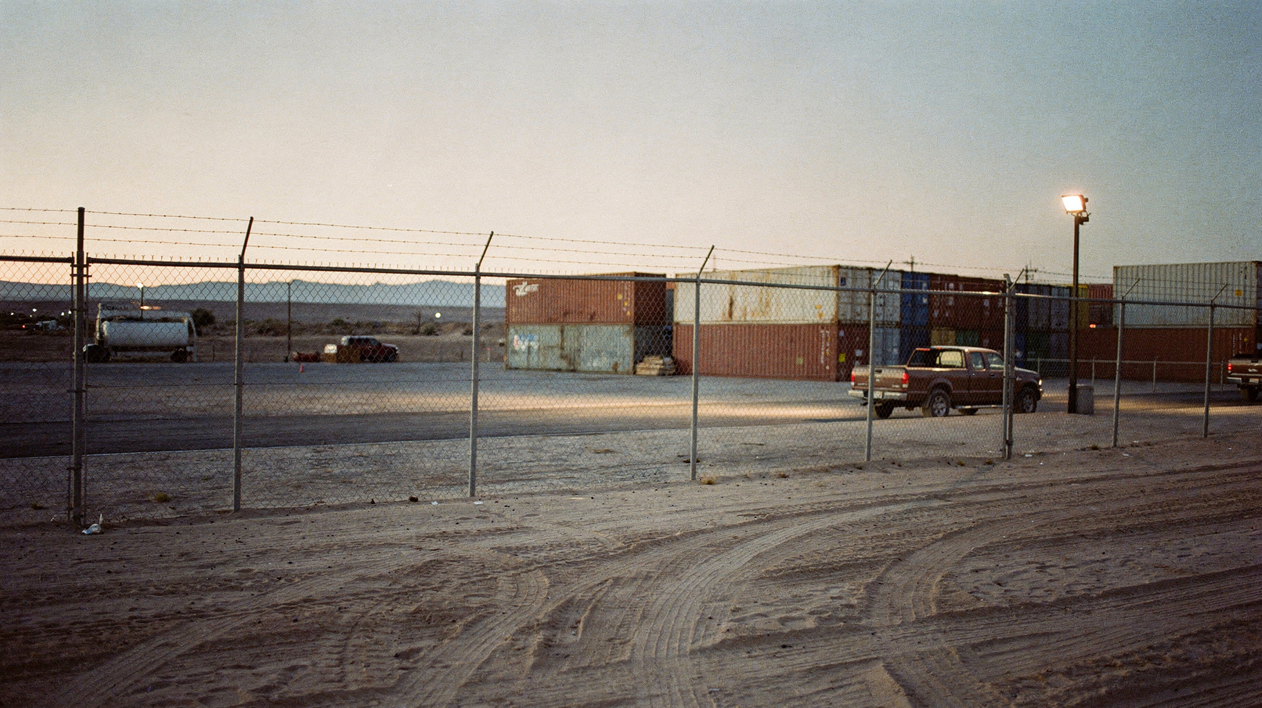 A tall chain-link fence stretching across a desert lot at dusk, shipping containers stacked behind it, a single floodlight on a pole, tire tracks in the sand