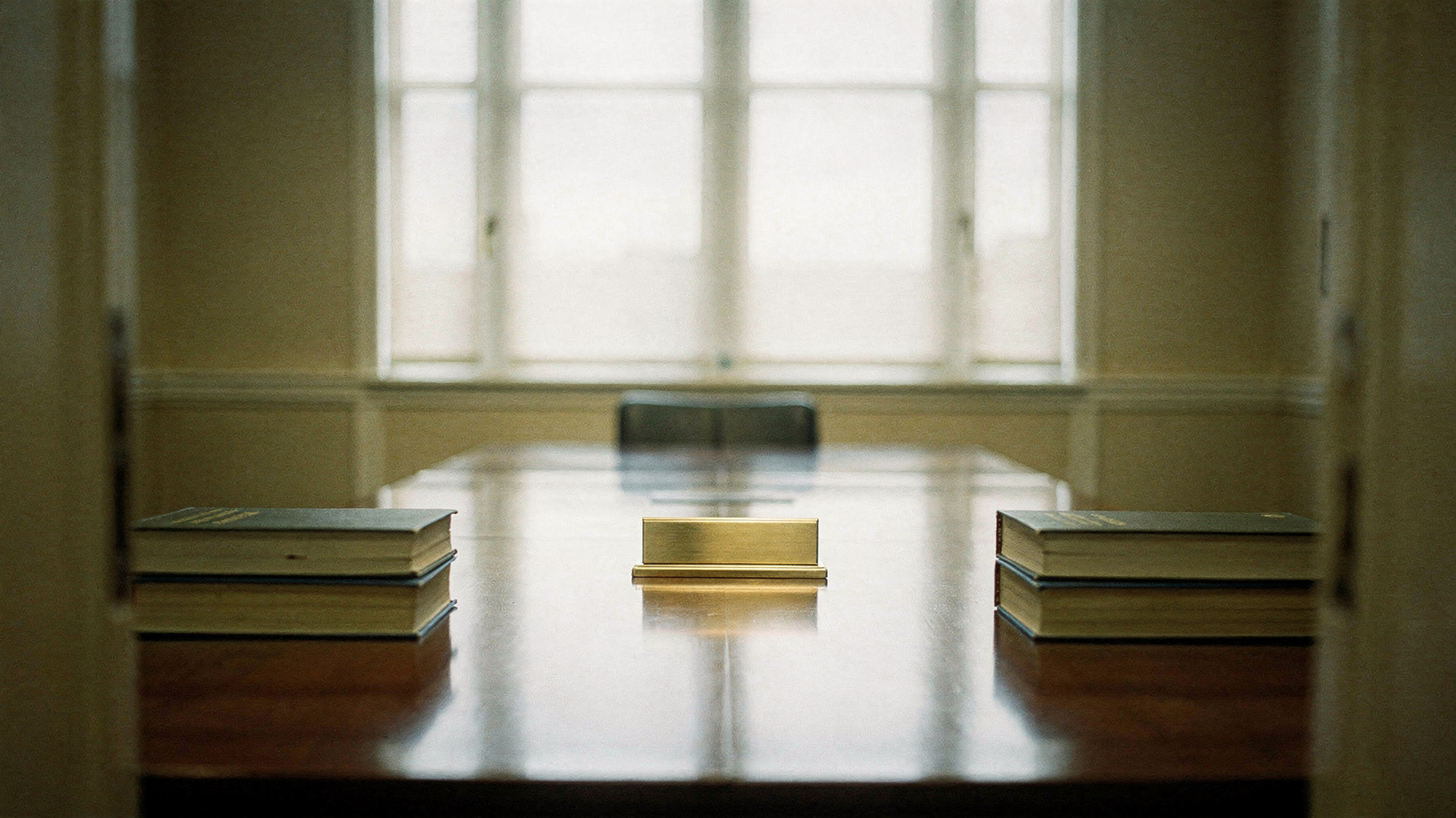 A documentary photograph of a federal agency conference room, two thick bound document volumes stacked at each end of a long polished table, an empty brass nameplate holder at the head, tall institutional windows behind