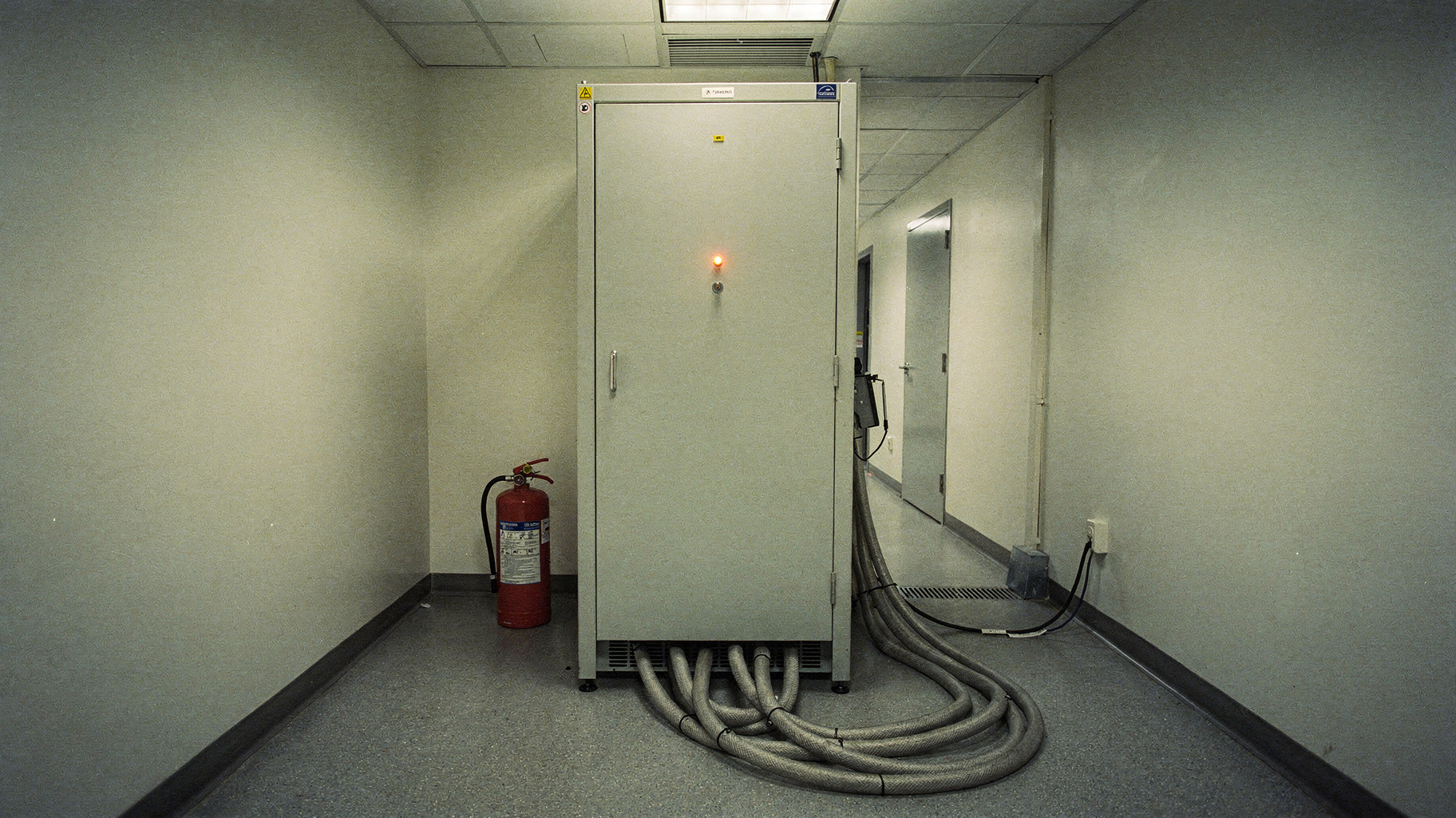 A documentary photograph of a sealed instrumentation cabinet in a laboratory corridor, a single amber status light glowing on its front panel, thick braided cables running from its base into the wall, polished concrete floor
