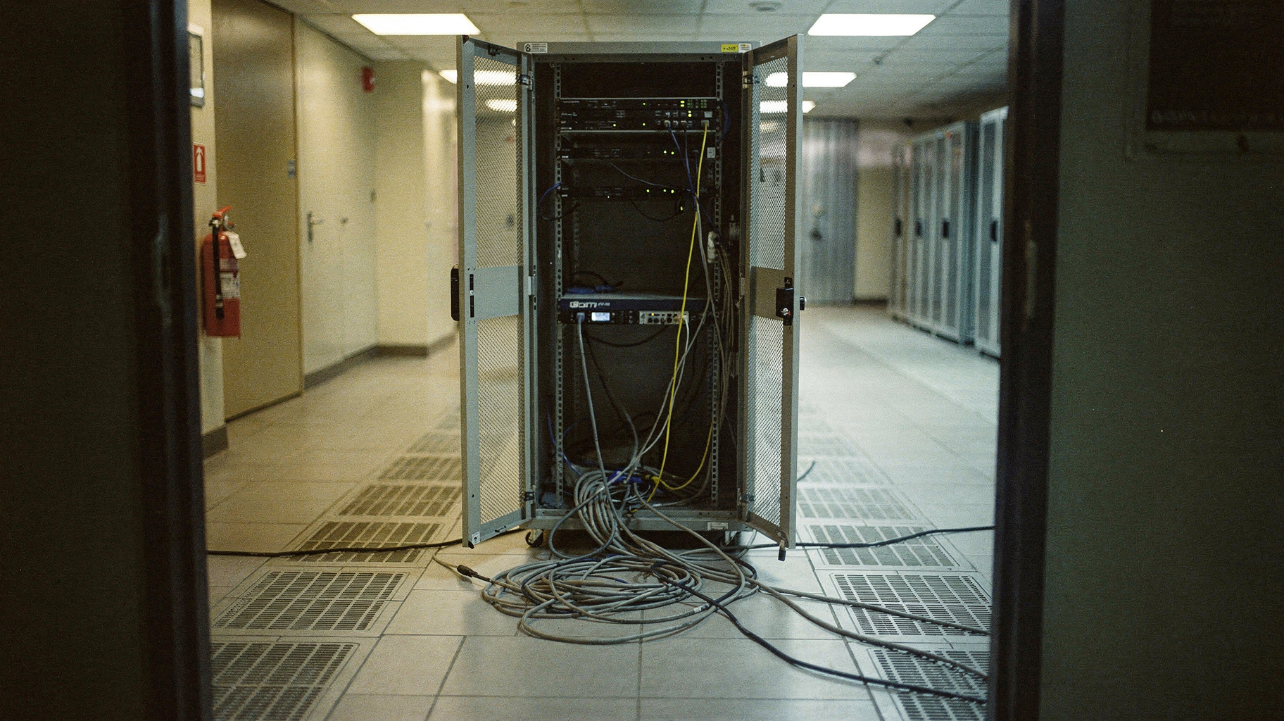An empty server cage with its door ajar and a cable run draped across the floor of a sparse data room