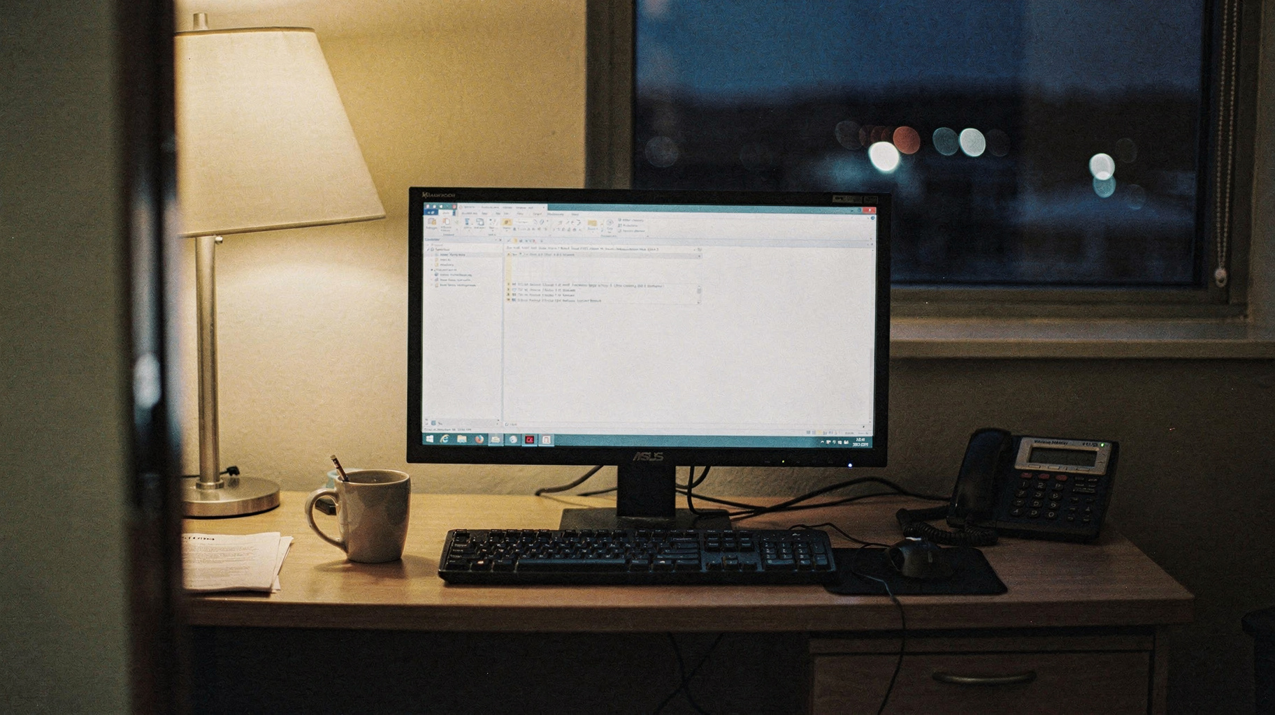 A documentary photograph of a desktop monitor and keyboard on an office desk at night, lit by a single desk lamp, a coffee mug and desk phone beside the keyboard