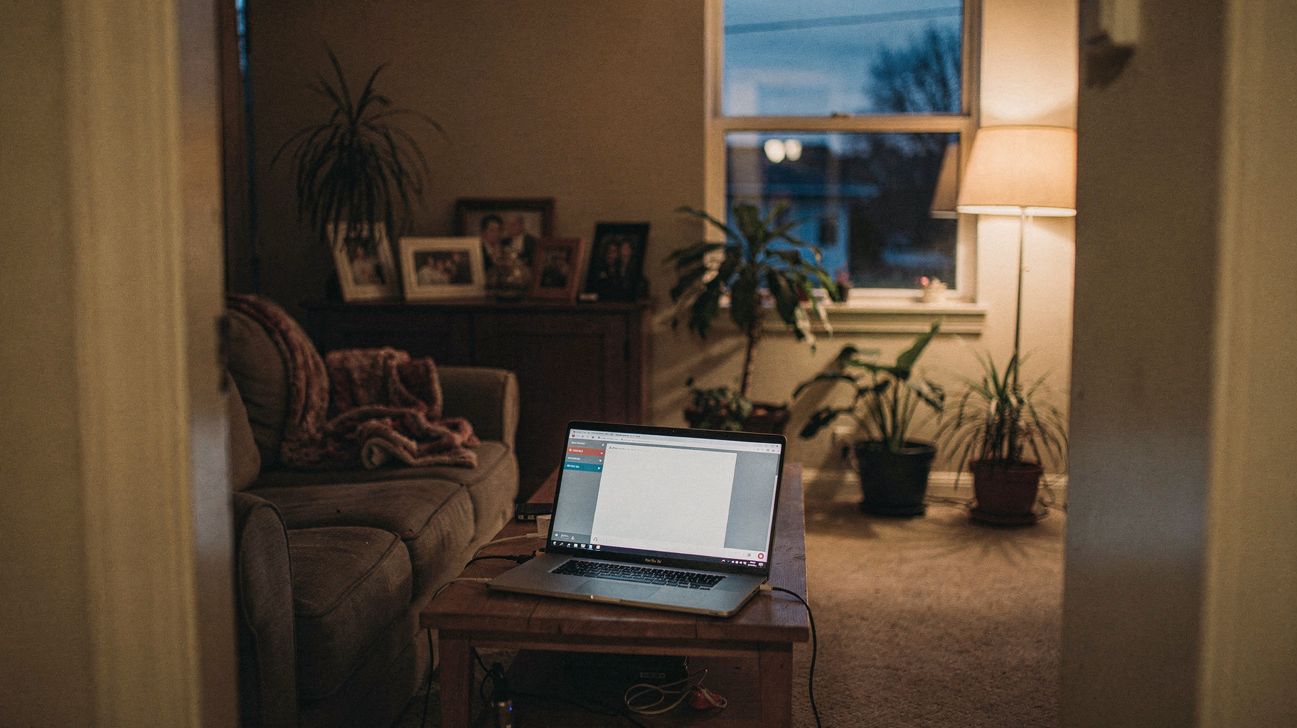 A documentary photograph of a suburban living room in the evening, laptop open on a coffee table, lamp light and TV glow mixing, family photos on the wall