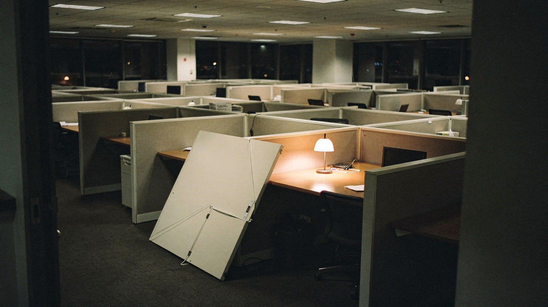 A vast empty corporate office floor at night, one white partition knocked over and lying on its side, a single desk lamp still on, muted earth tones on film