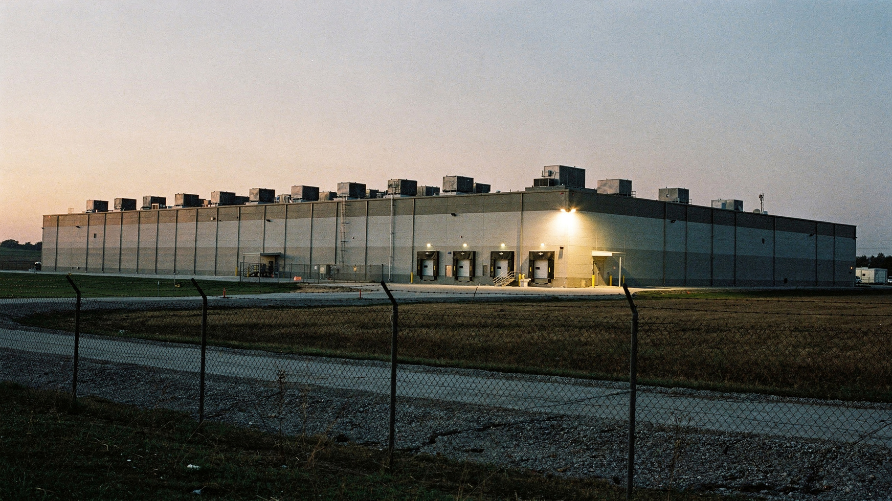 A documentary photograph of a long low data-center building on flat Ohio farmland at dusk, rows of cooling units along its roofline, a single floodlight glowing at the loading dock, wide empty sky above