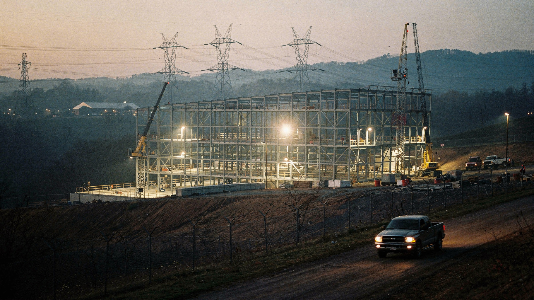 A vast data center construction site at dusk, transmission towers receding into hazy hills, work lights on steel framing, muted earth tones on film