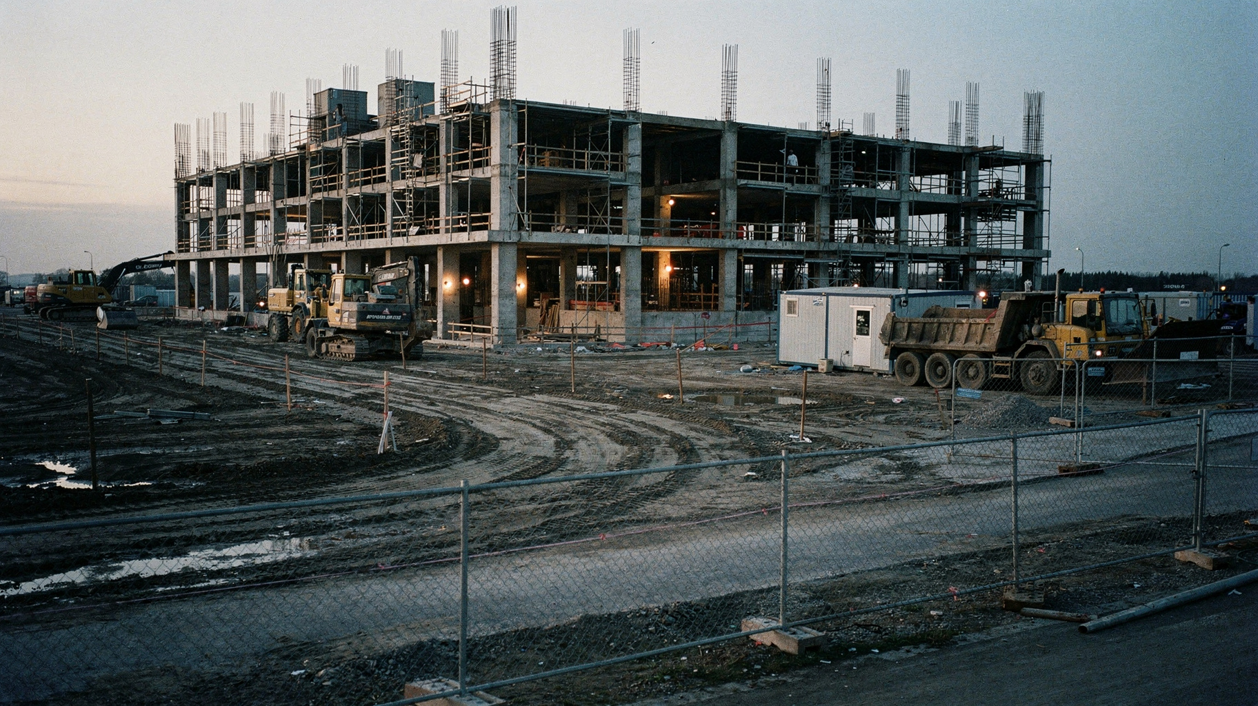 A documentary photograph of a construction site at dawn beside a half-built data center shell, rebar and scaffolding silhouetted against first light