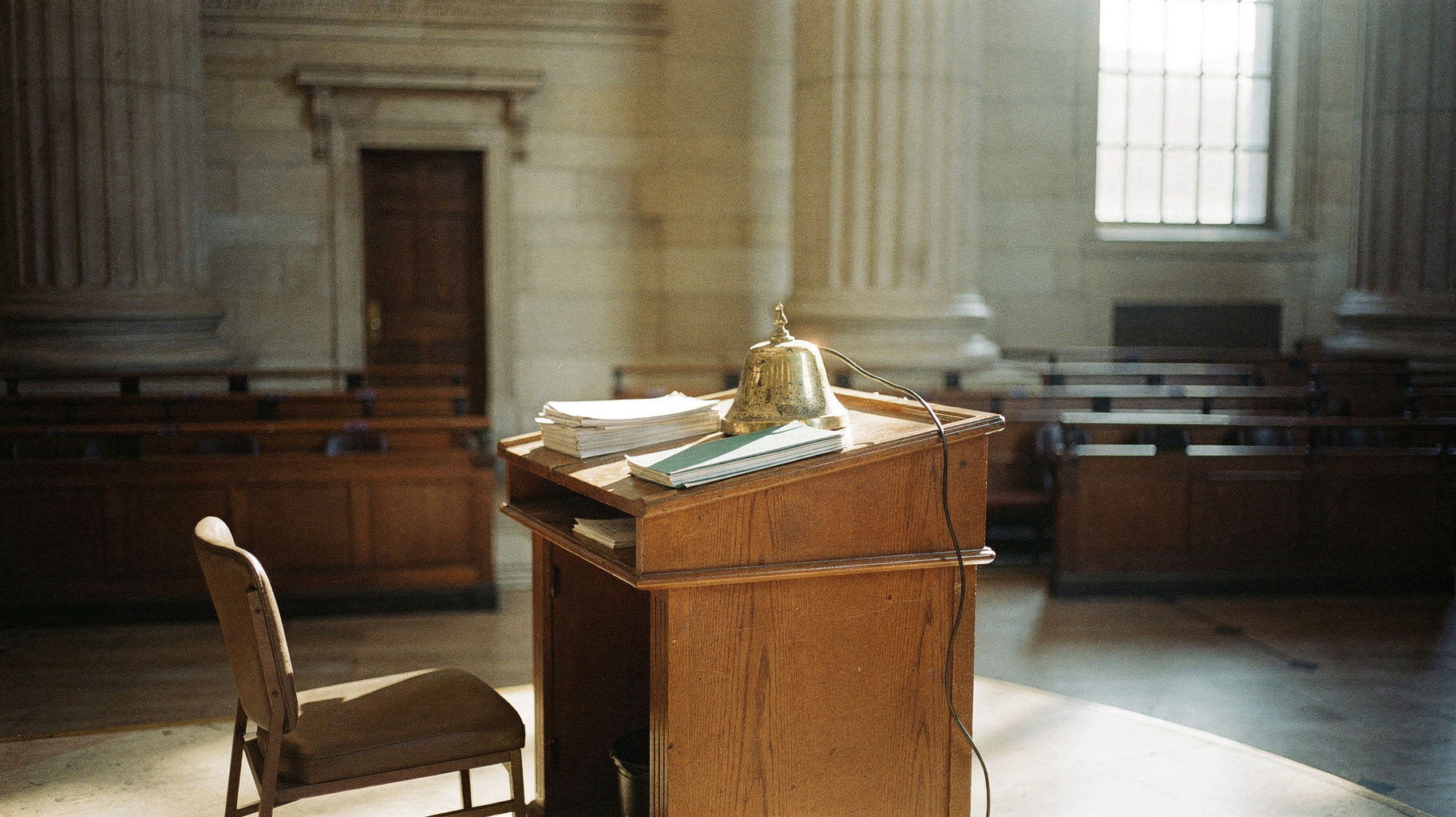A documentary photograph of an empty stock exchange podium with a brass opening bell, morning light through tall windows, stacked papers on the lectern, nobody in frame