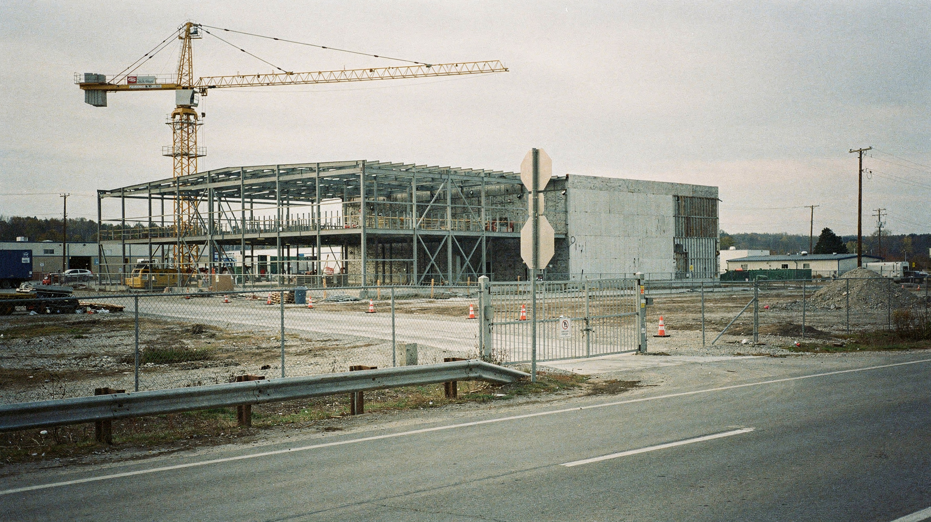 A half-finished data center shell, open steel frame with a crane standing idle beside it, seen from across a fenced gravel lot under flat overcast light