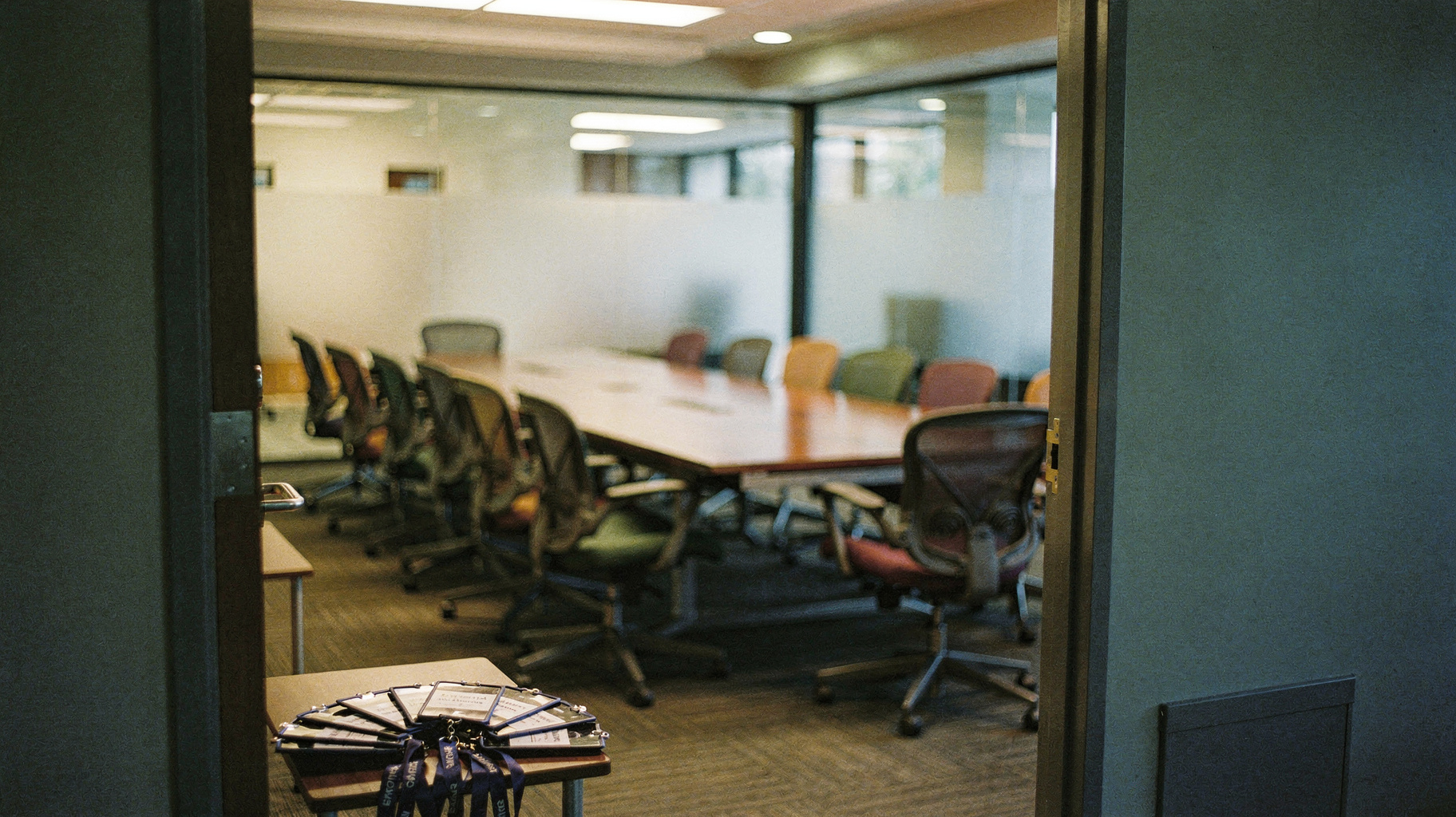 A documentary photograph of an empty conference room in early morning, long table with mismatched office chairs from different companies, visitor badges fanned on a tray by the door, nobody in frame