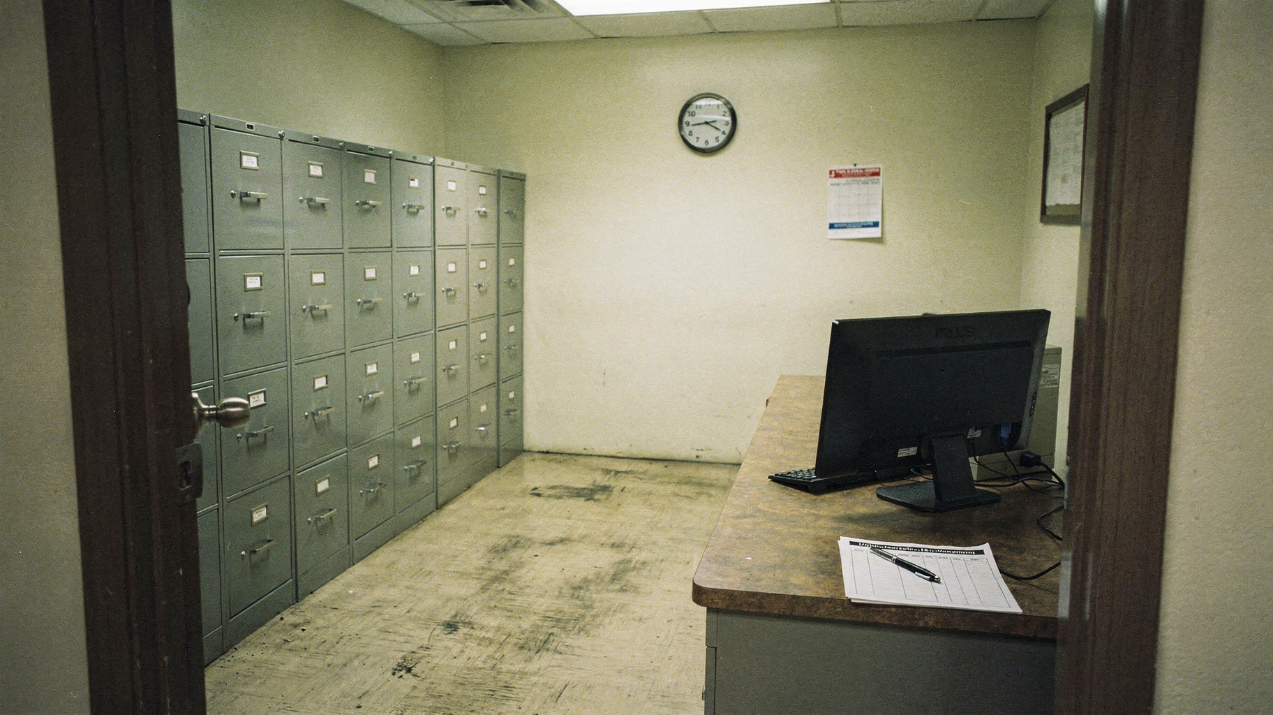 A government records room photographed from the doorway, gray metal filing cabinets along one wall, a single desk with a switched-off monitor and a visitor sign-in sheet, hard fluorescent light, no people, documentary distance, muted institutional tones, visible film grain