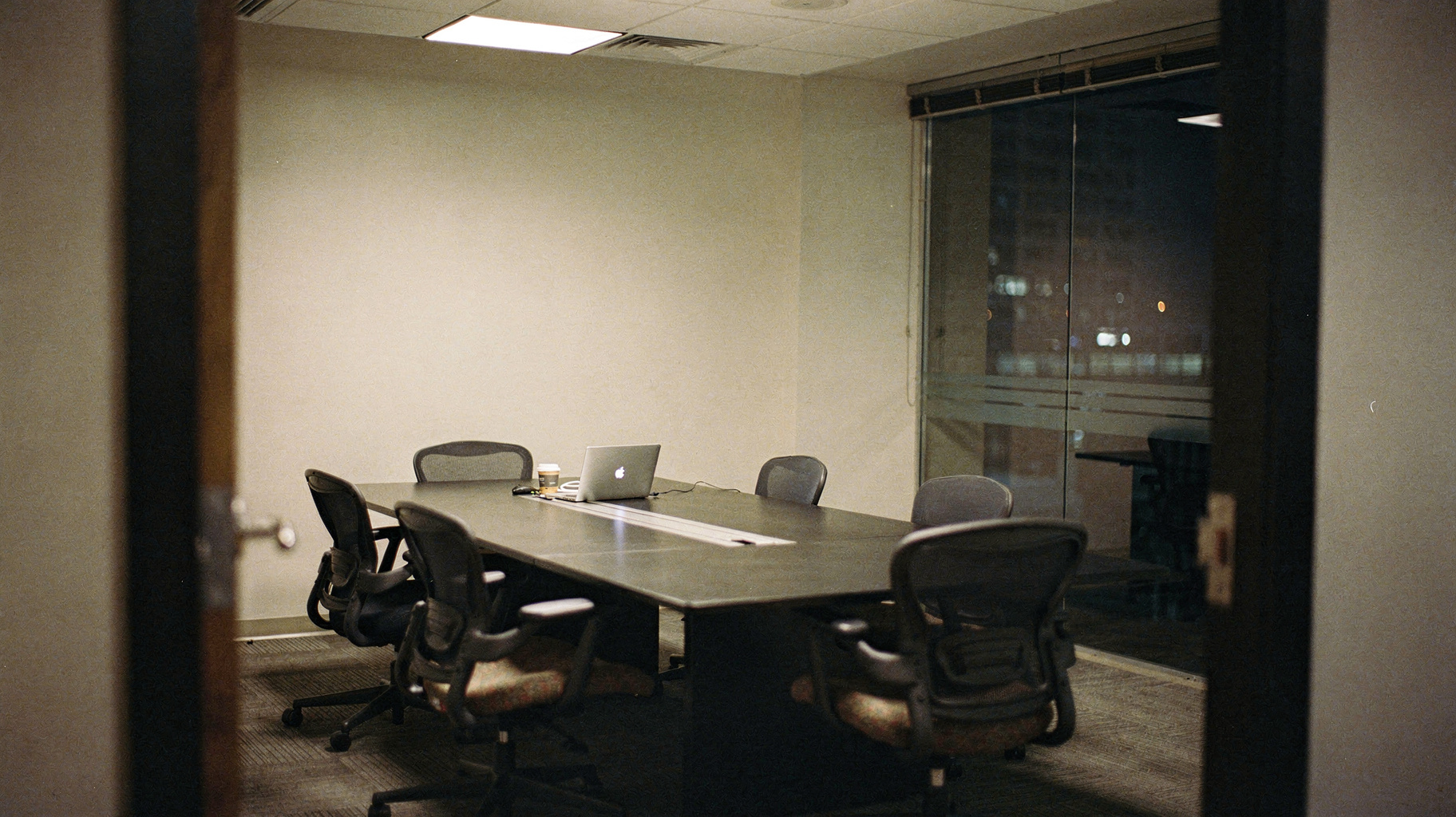 A documentary photograph of an emptied conference room at night, mismatched chairs pushed back from a long table, one abandoned laptop and a half-finished cup of coffee, blinds half drawn