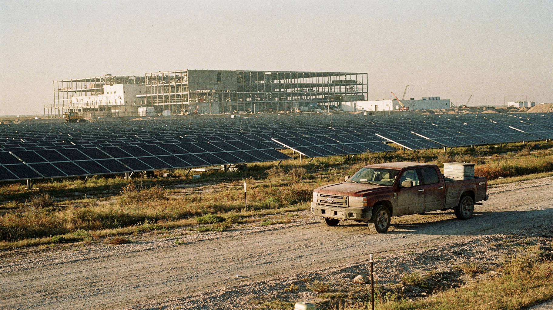 A vast solar array stretching toward a half-built data center shell, a pickup truck on a gravel service road, early morning haze