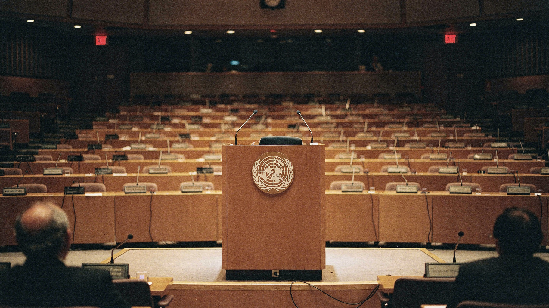 An empty lectern with the United Nations emblem in a large assembly hall, rows of empty delegate seats receding behind it, house lights low, photographed from an observer's seat at a documentary distance