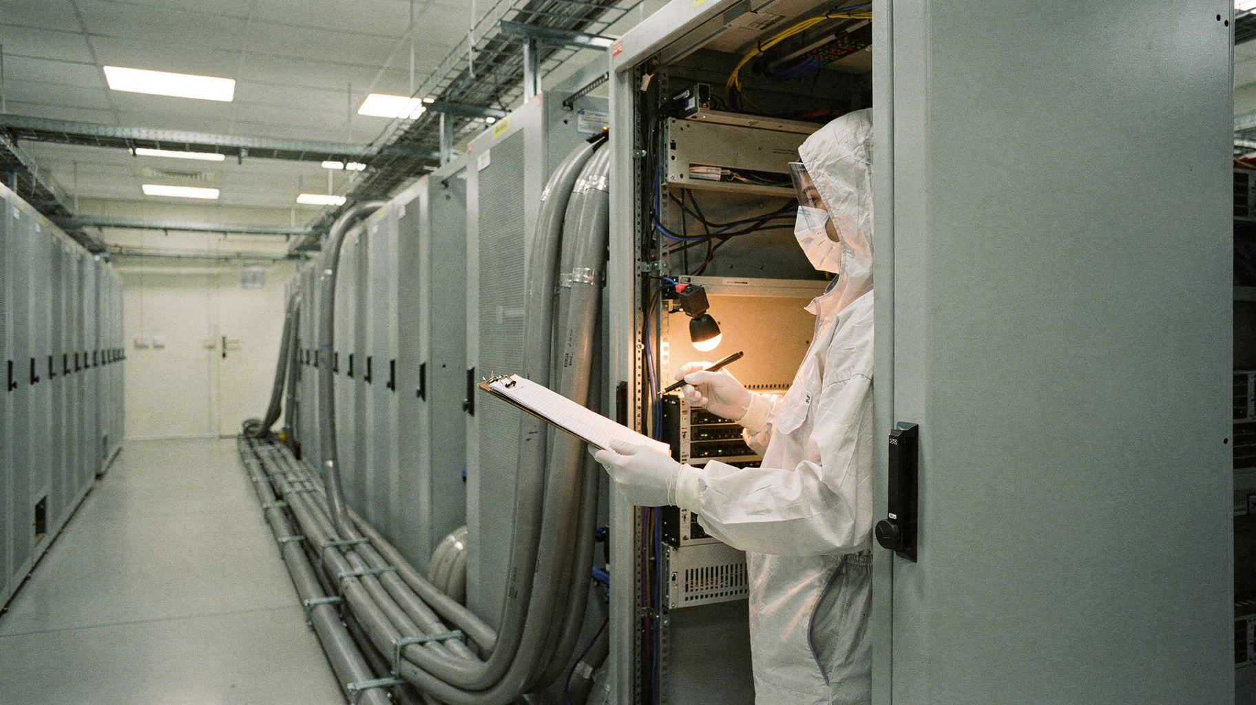 A cleanroom technician in a white bunny suit inspecting a server rack with thick coolant lines running into it, clipboard in hand, another rack receding down the aisle