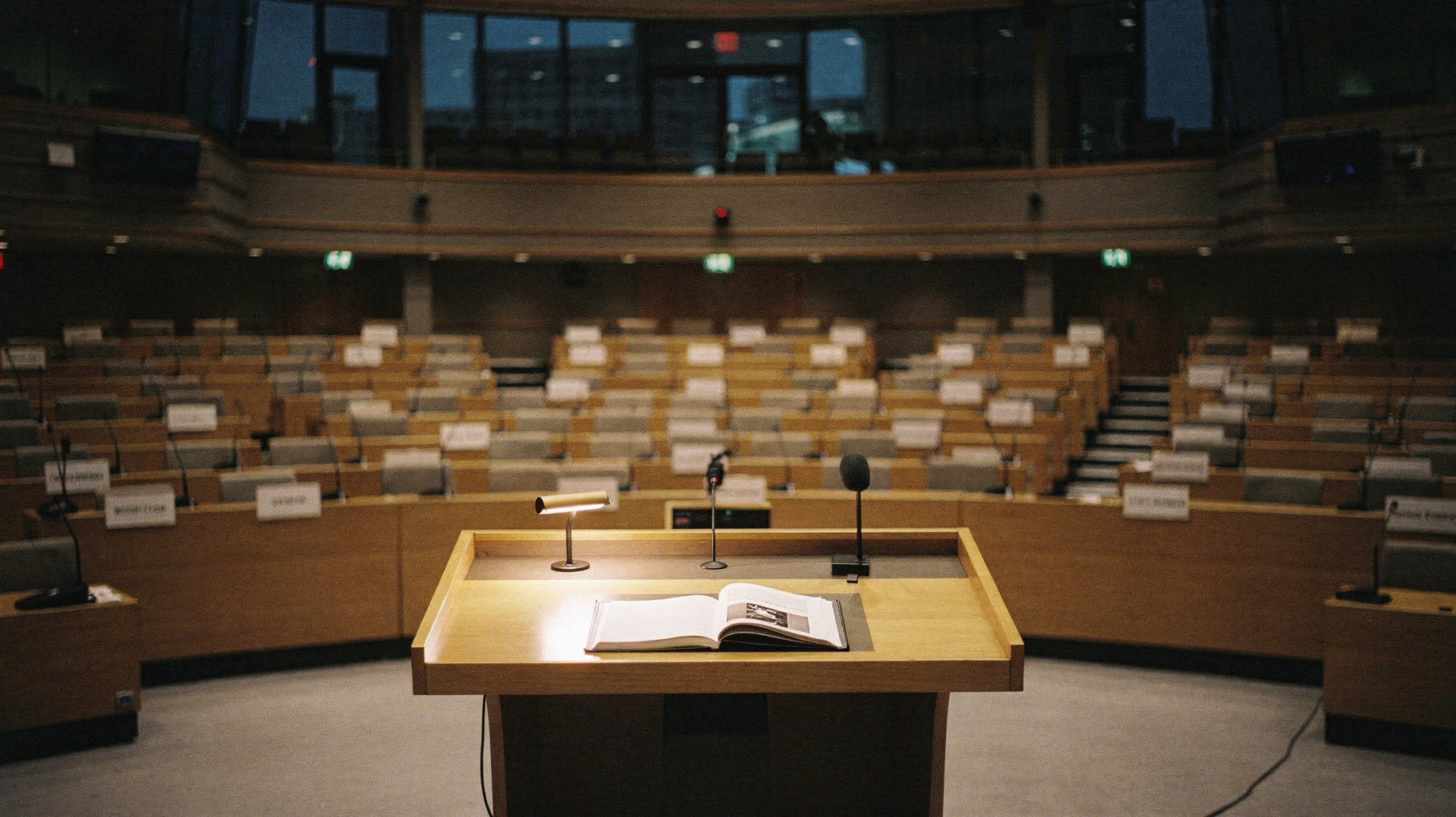 A documentary photograph of a near-empty international conference chamber, curved rows of delegate desks with blank placards, a single bound report open on the central lectern, tall windows dimmed behind