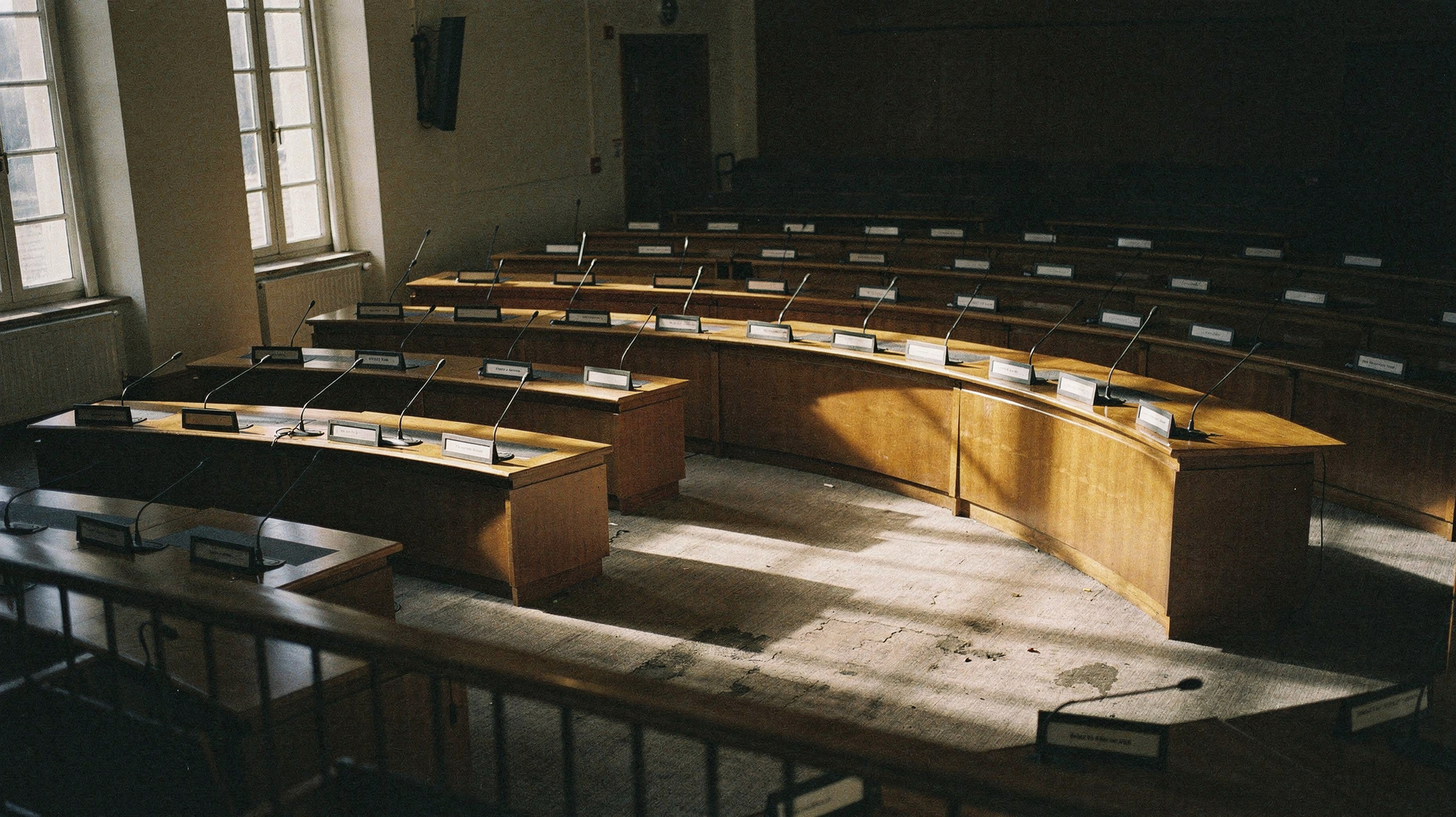 A near-empty council chamber photographed from the public gallery, rows of curved wooden desks with microphones and nameplates, one tall window casting hard afternoon light across the floor, documentary distance, no people visible, muted earth tones, visible film grain